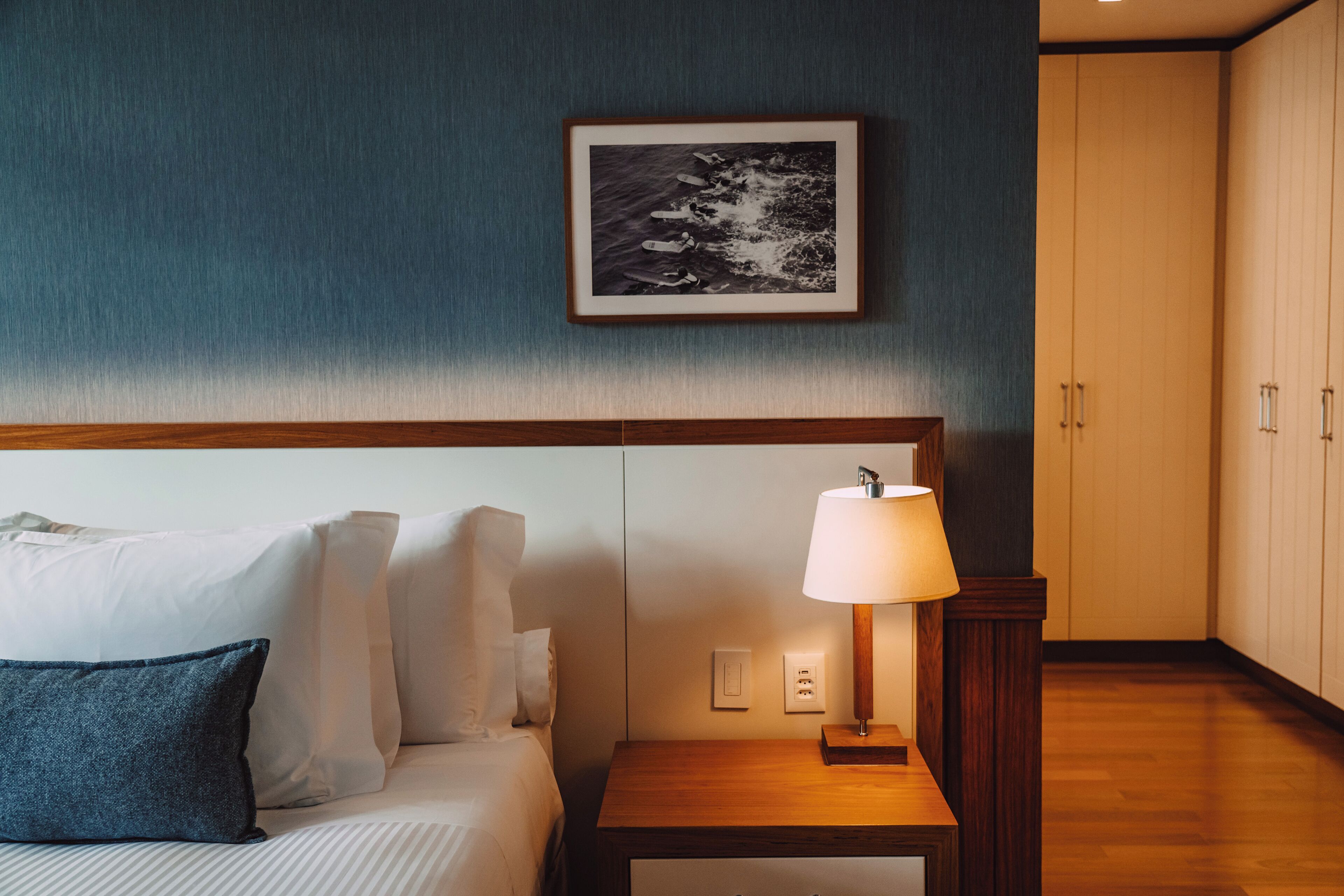 A hotel room with a bed, pillows, a bedside table with a lamp, a framed picture on the wall, and a wardrobe.
