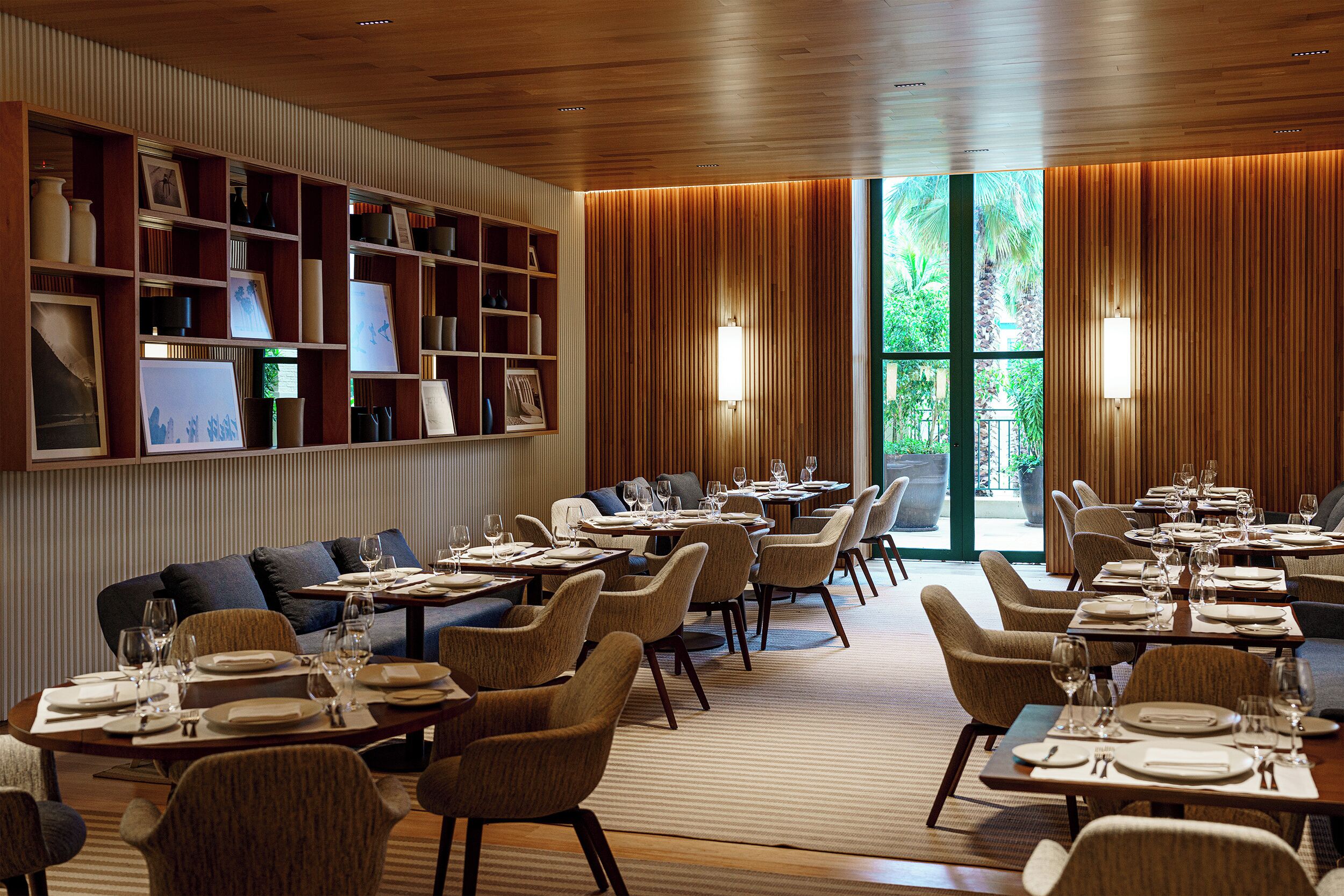 Restaurante com teto de madeira, prateleiras com quadros e vista para o verde através de amplas janelas.