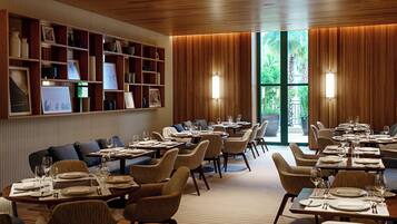Restaurante com teto de madeira, prateleiras com quadros e vista para o verde através de amplas janelas.