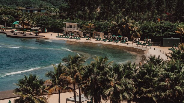 Un complejo turístico en la playa con zona de piscina, sillones de descanso y una torre de socorristas.