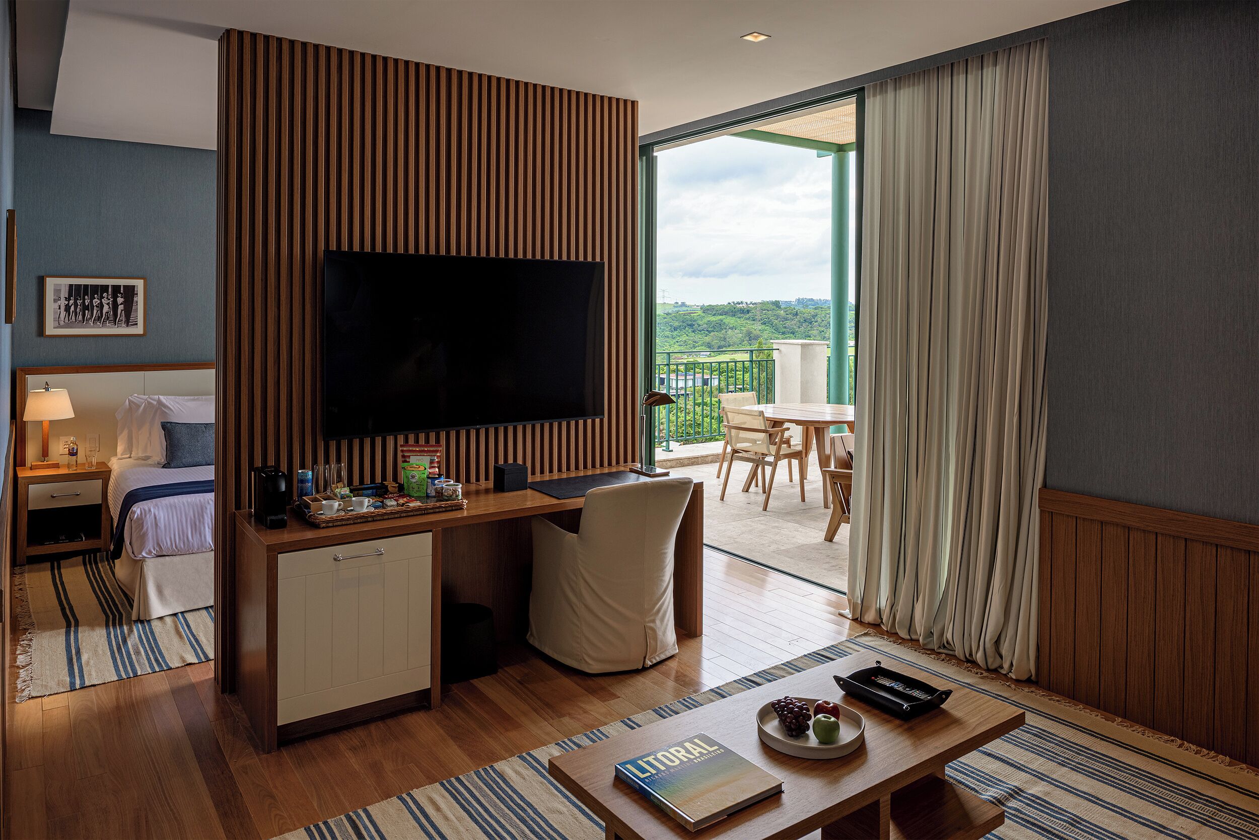 A hotel room with a TV, a desk, a bed, a balcony with a view, and a dining area.