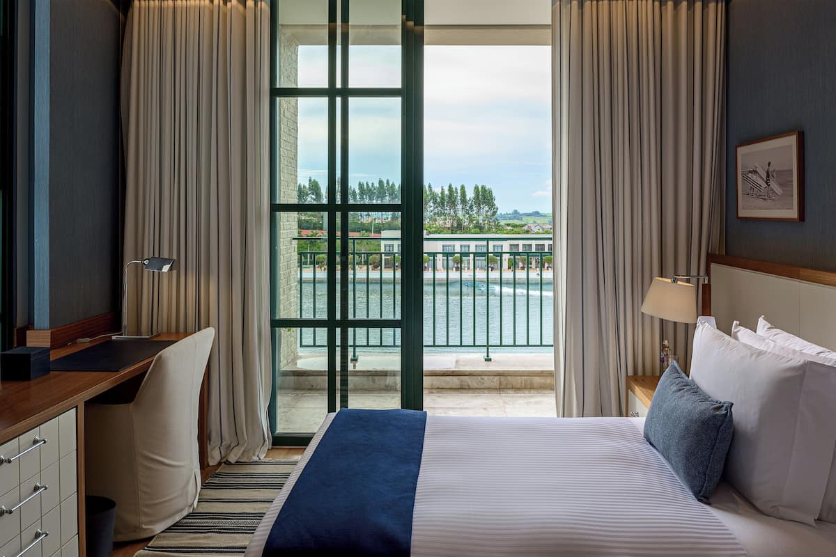 Hotelkamer met een bed, bureau en stoel, uitzicht op water en gebouwen, en een raam met gordijnen.
