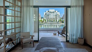 Habitación de hotel con una cama grande, una zona de estar con sillones y vistas a un edificio y una piscina a través de las puertas del balcón.