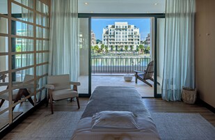 Quarto de hotel com uma cama grande, área de estar com cadeiras, vista de um edifício e piscina através das portas da varanda.