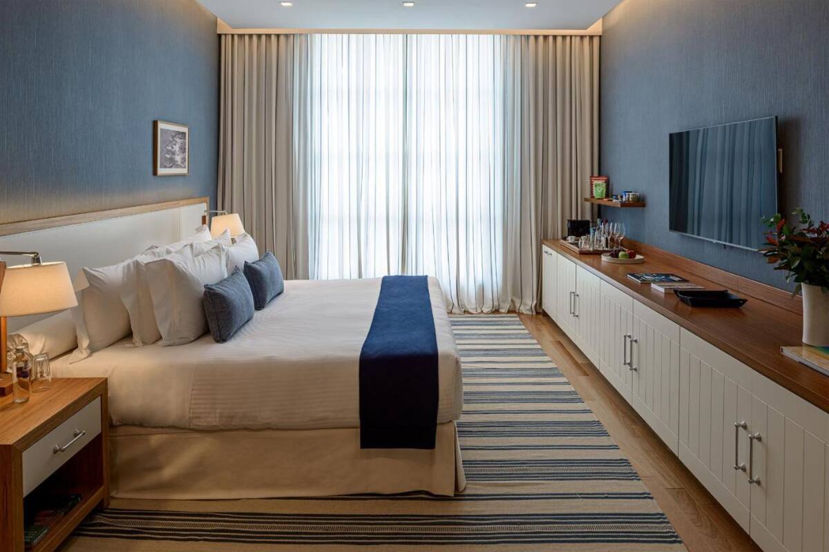 Een hotelkamer met een bed, nachtkastje, televisie en een groot raam met gordijnen.