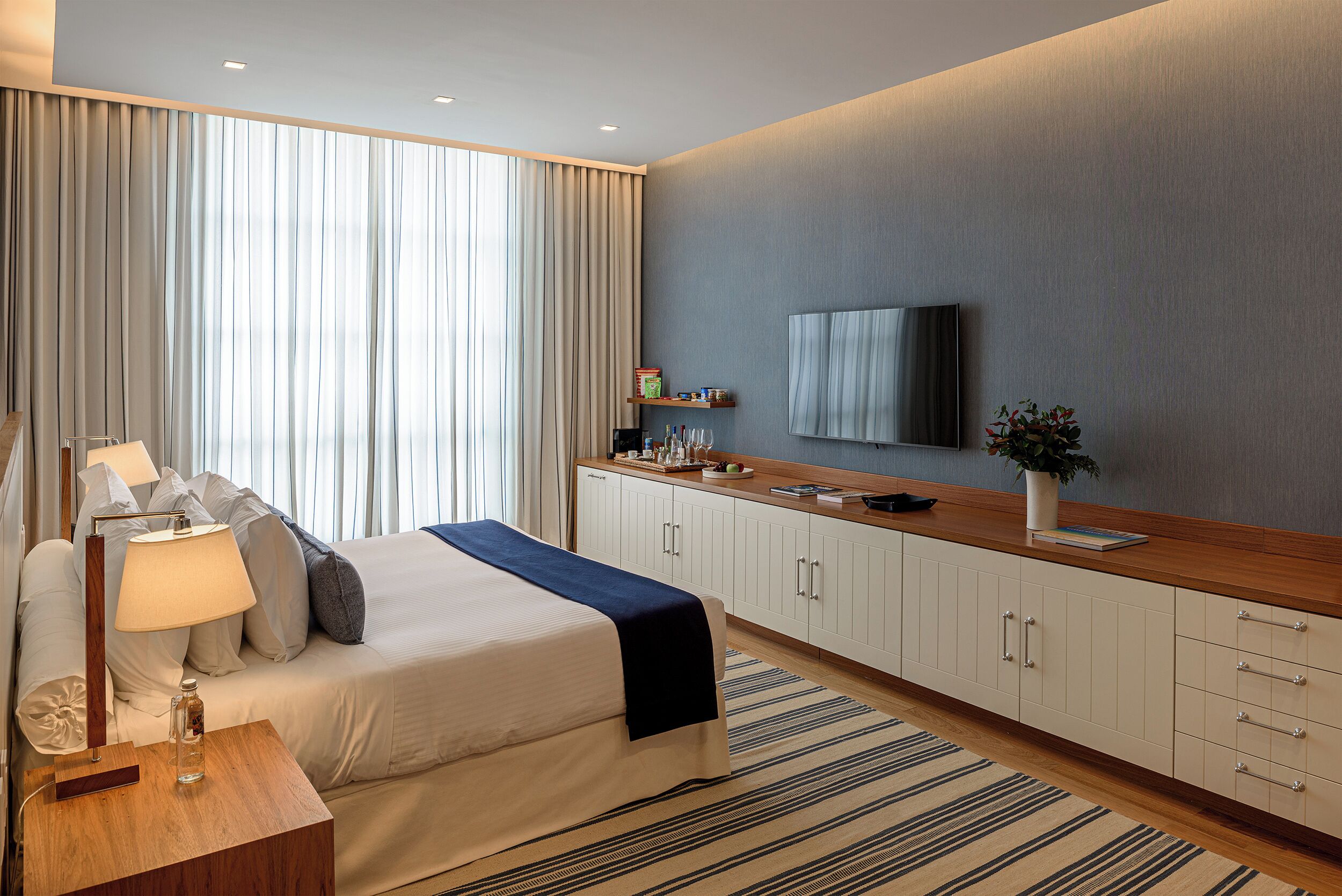 A modern hotel room with a large bed, a bedside table with a lamp, a TV mounted on the wall, and a small shelf with toiletries.
