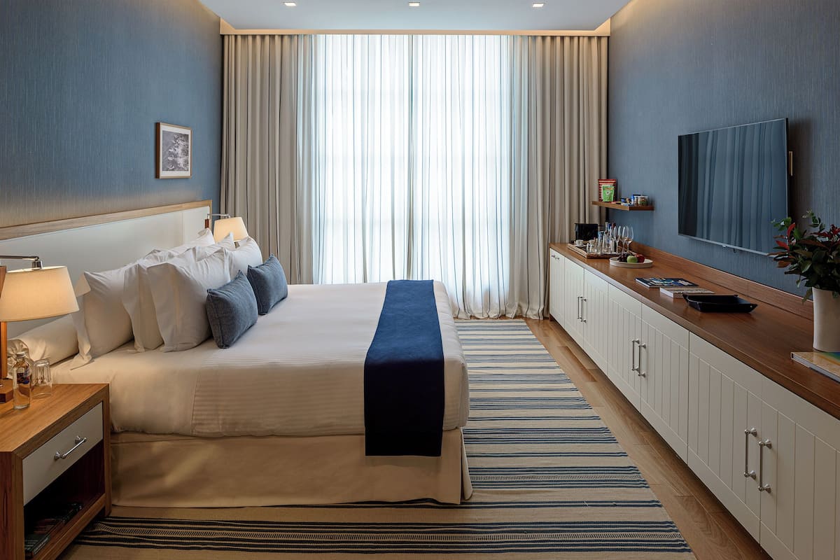 Een hotelkamer met een groot bed, een nachtkastje met een lamp, een aan de muur bevestigde televisie en een raam met gordijnen.