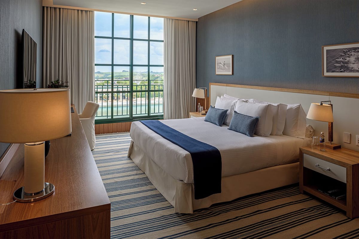 Hotelkamer met groot bed, nachtlampjes, nachtkastje van hout, uitzicht op de omgeving door een raam en een ingelijst schilderij aan de muur.