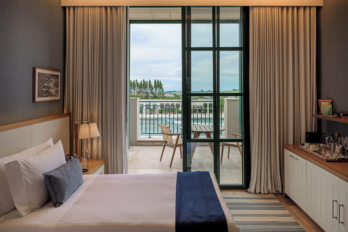 Een hotelkamer met een groot bed, een bureau met lamp, uitzicht op het zwembad en terrasmeubilair, en een ingelijst schilderij aan de muur.