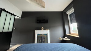 A bedroom with a bed, a hanging light fixture, a fireplace, and a window with blinds. - Aux Deux Tours (Leval)