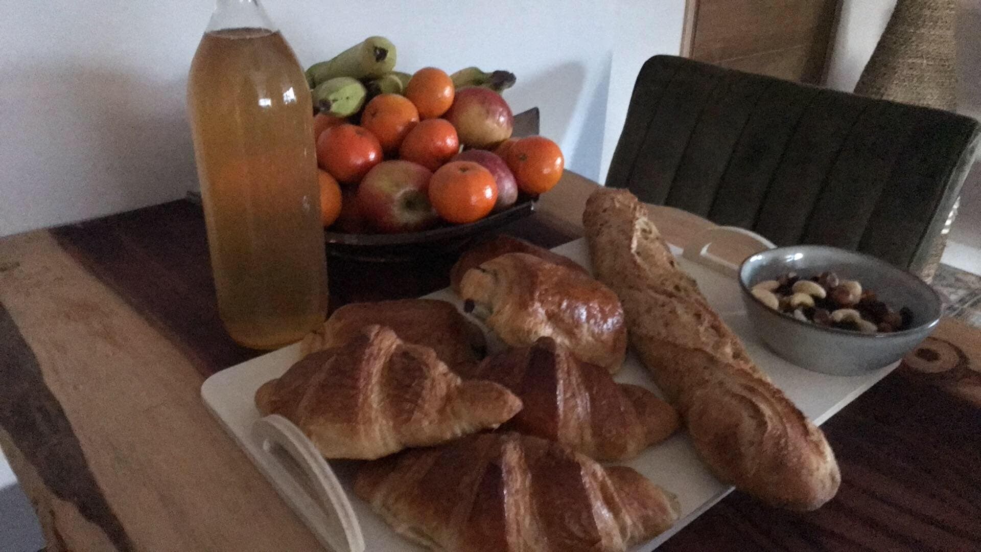 Une table avec une bouteille, un saladier de fruits, une assiette de croissants et un bol de noix.