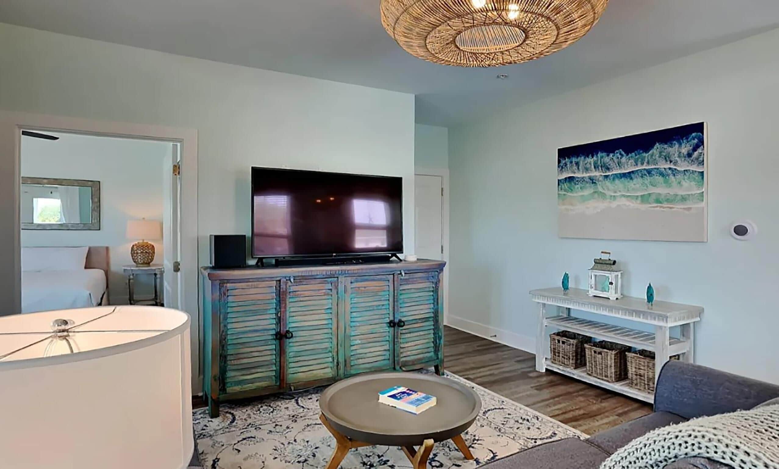 A living room with a television on a cabinet, a round coffee table with a book, a wicker ceiling light, and a painting of waves on the wall.