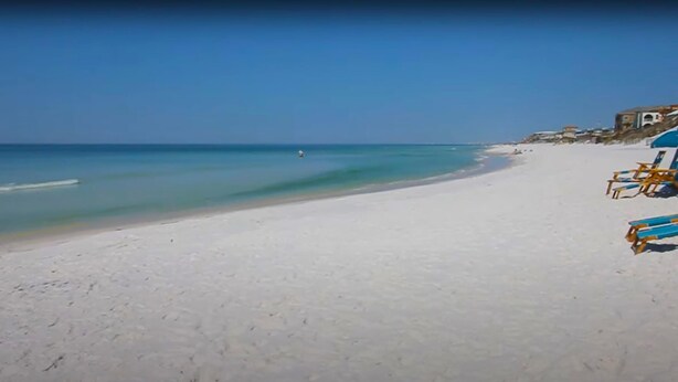 Une plage de sable blanc bordée d’une eau d’un bleu cristallin. On y trouve quelques chaises de plage et des parasols, situés sur le côté droit.