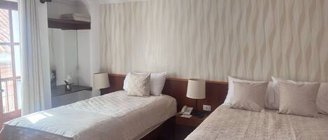 Habitación de hotel con dos camas, un ventanal con cortinas, un espejo, una mesita de noche con lámpara y una pared con un diseño ondulado.