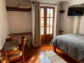 Una habitación con cama, escritorio y silla, ventana con cortinas y televisión montada en la pared.