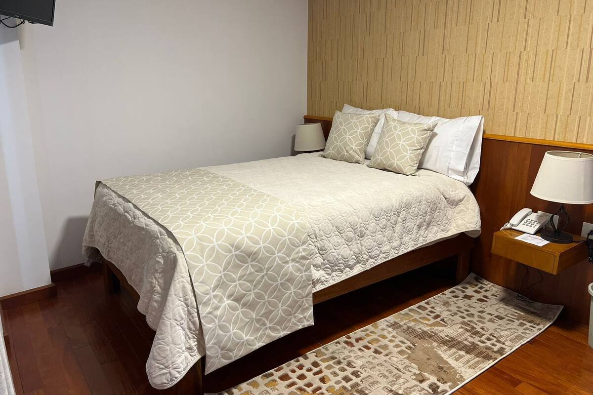 Una cama bien tendida con una colcha y almohadas estampadas, una mesita de noche con una lámpara, y una alfombra con diseño sobre piso de madera.