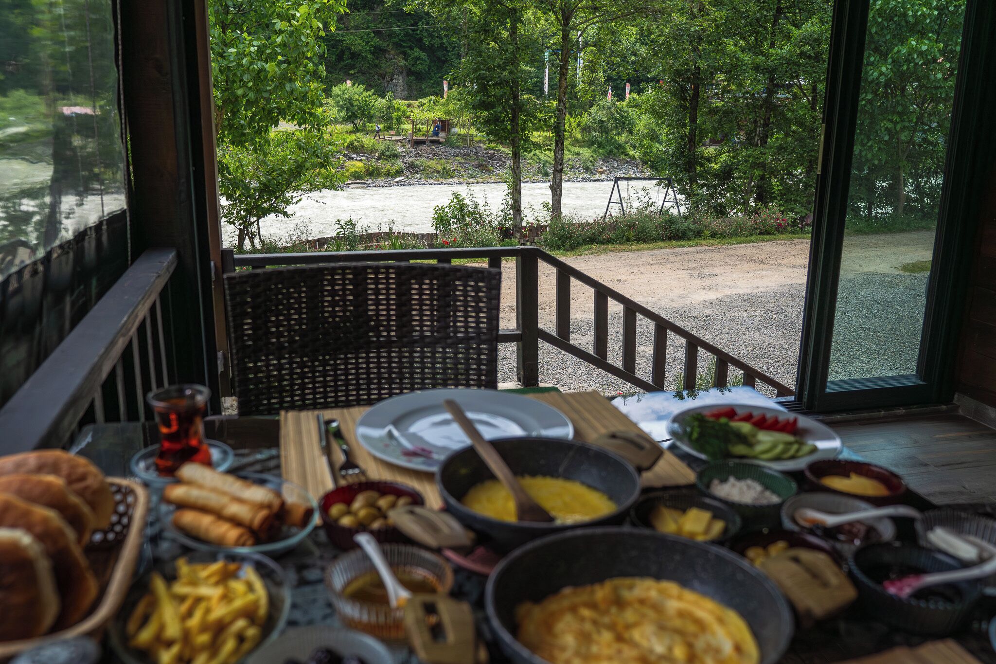 A dining table with various dishes and a view of a river and forest.