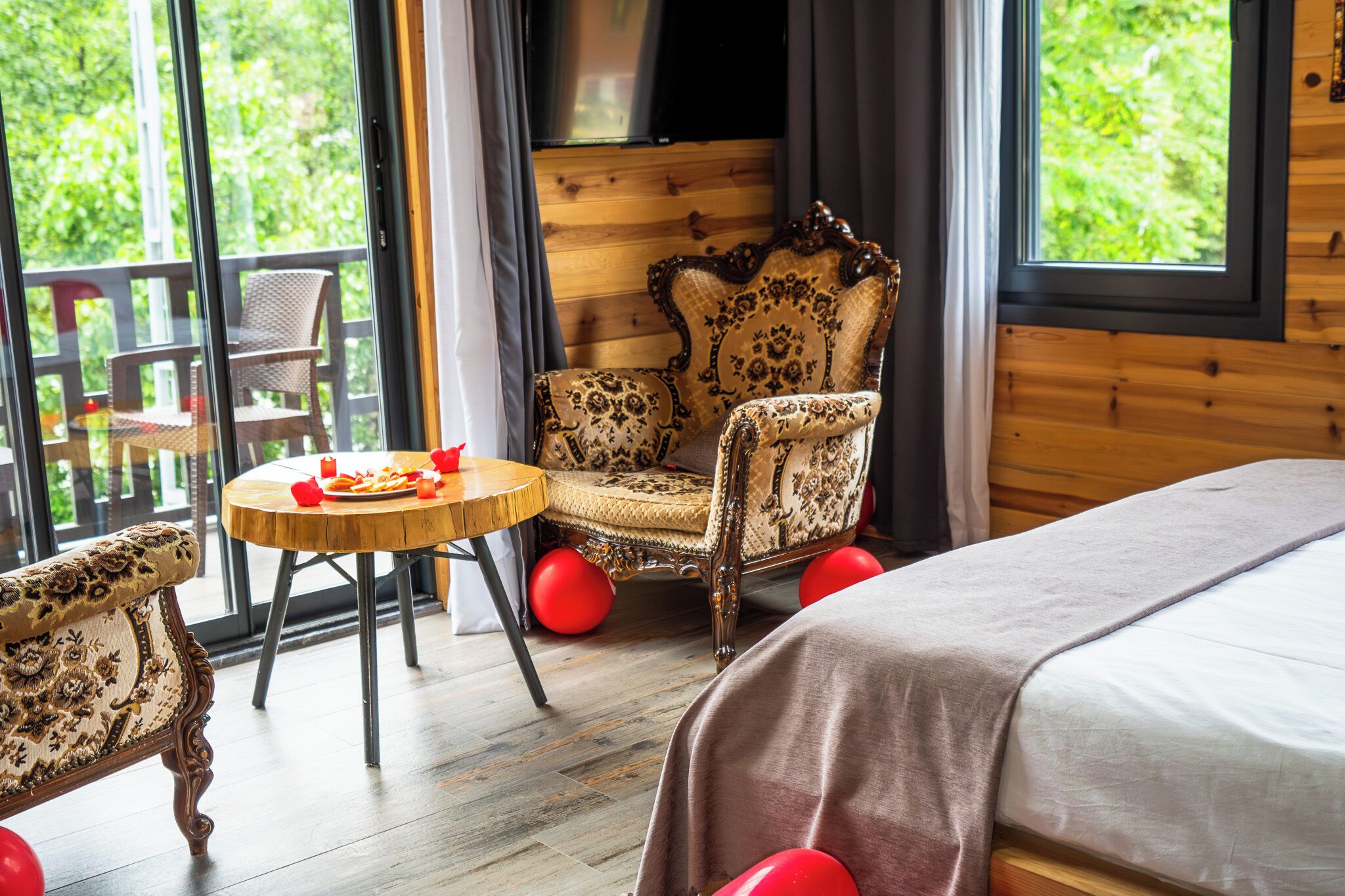 A room with a bed, a wooden chair, a round table with a red object, and a window with a view of greenery.