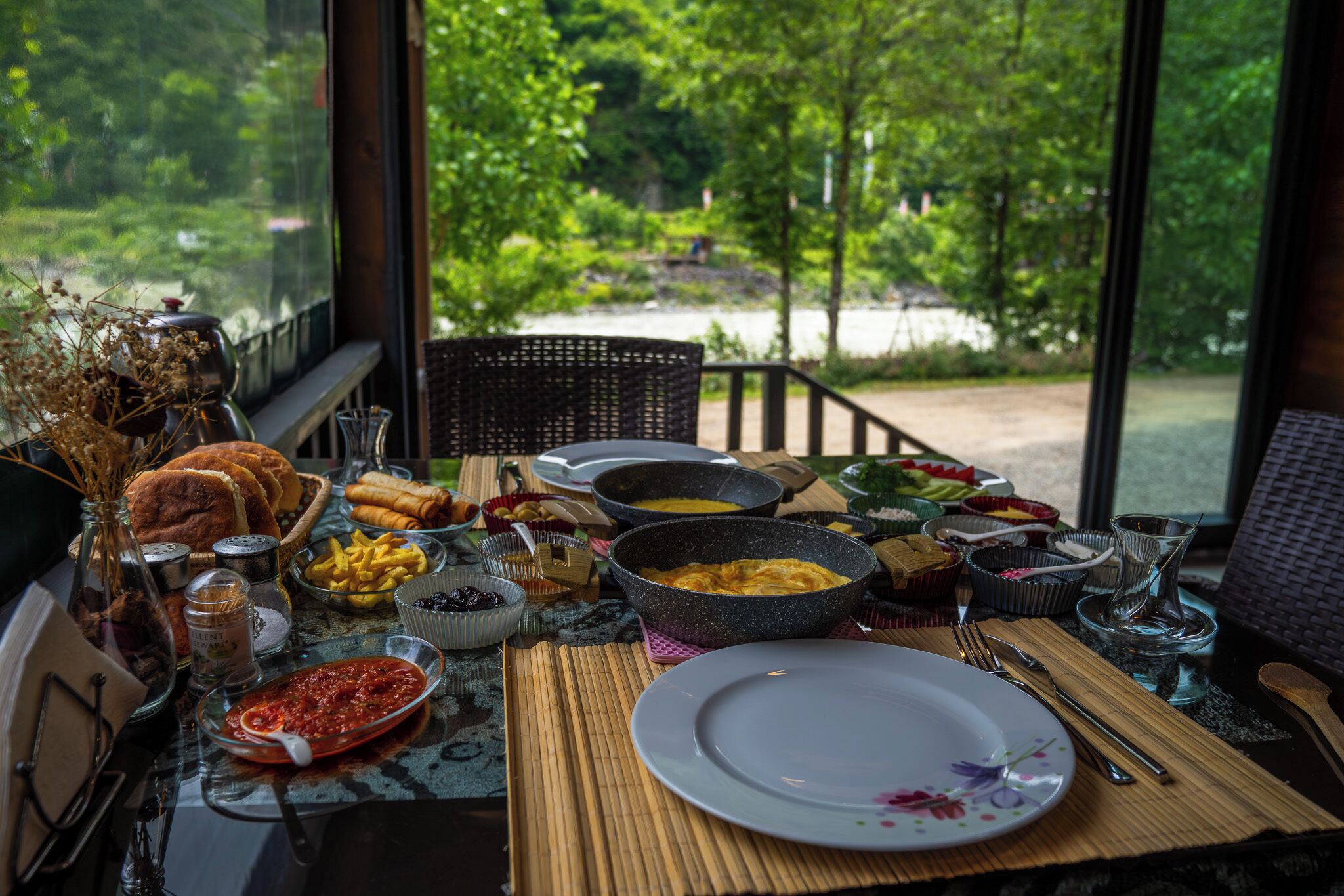 A dining table with various dishes, utensils, and a view of greenery outside.