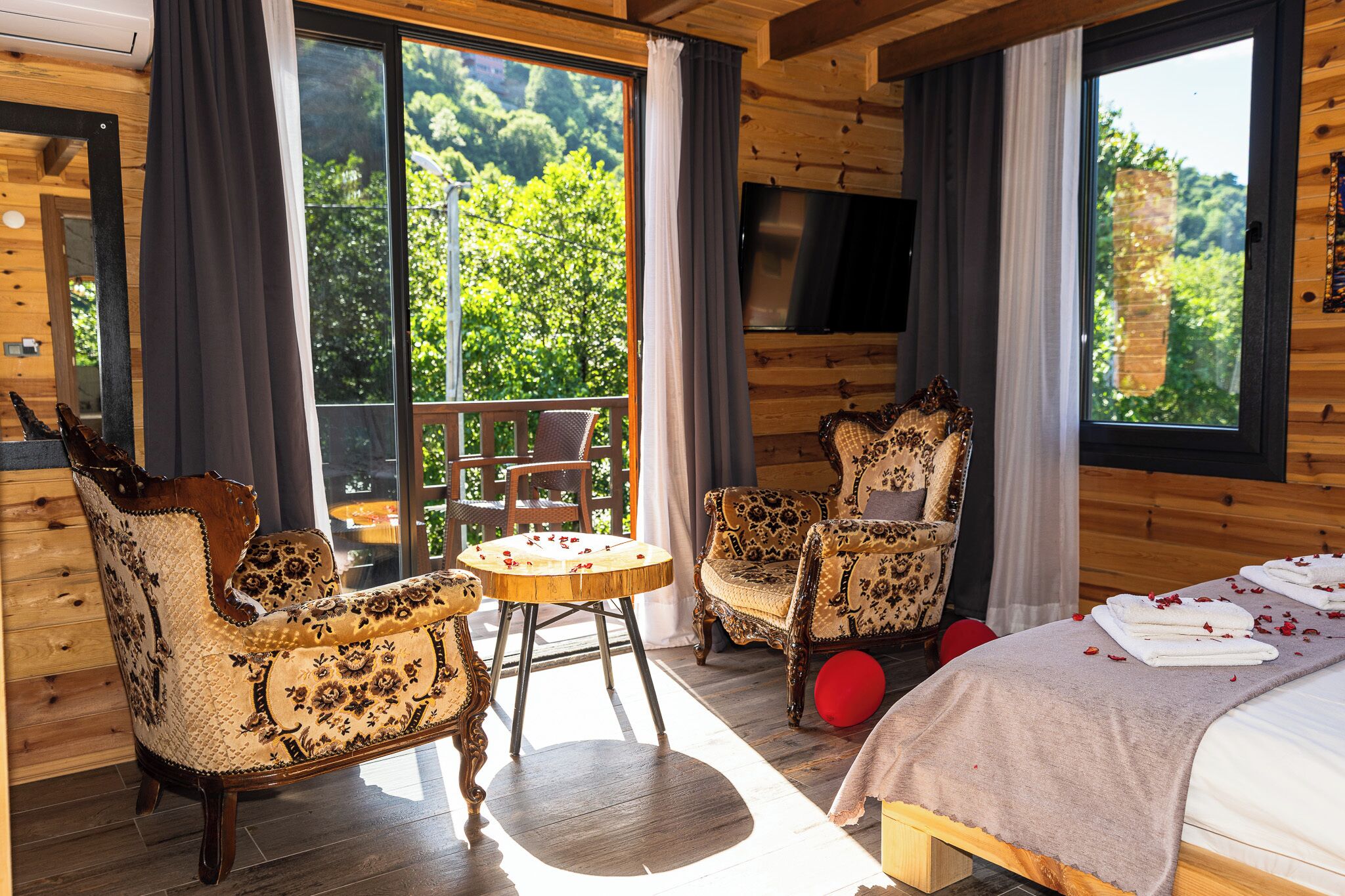 Une cabane en bois avec un balcon, deux fauteuils à motifs, une table ronde et un lit avec des serviettes.