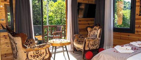 A wooden cabin with a balcony, two patterned armchairs, a round table, and a bed with towels.