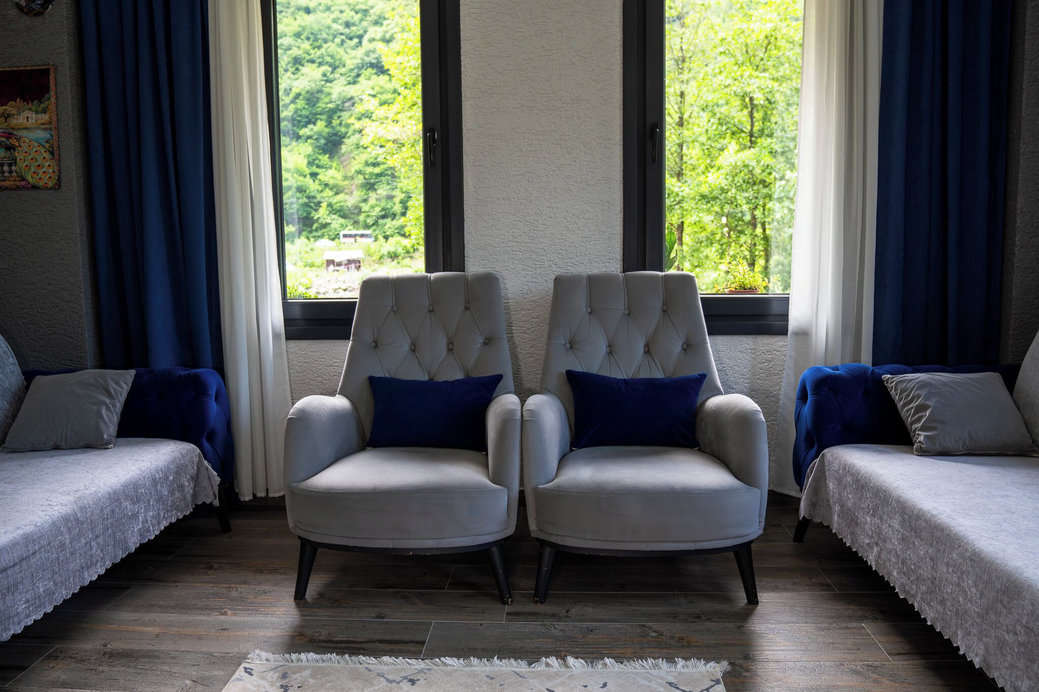 A room with two white armchairs, a sofa, and a view of greenery through large windows.