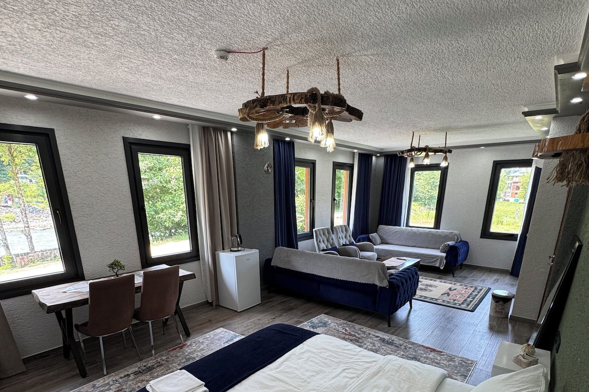 Moderni hotellihuone, jossa on suuri sänky, sohva, työpöytä ja tuoli. Huoneessa on suuret ikkunat ja kattotuuletin.