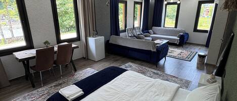 A modern hotel room with a large bed, a sofa, a desk, and a chair. There are large windows and a ceiling fan.