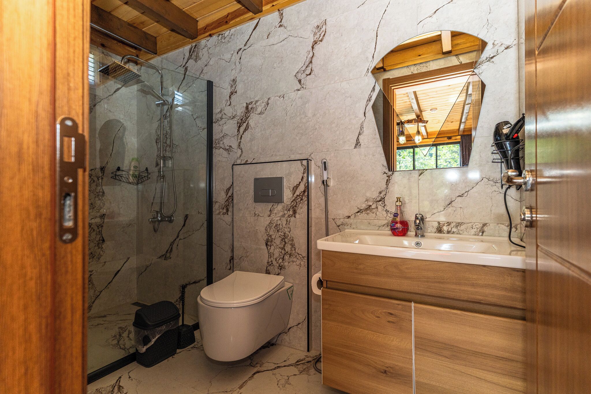 Un bagno con doccia vetro, un water, una toeletta in legno con specchio e pareti in marmo.