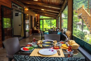 A dining area with a table set for a meal, a wicker chair, and a view of greenery outside. - Pallas Suit  (Ardeşen)