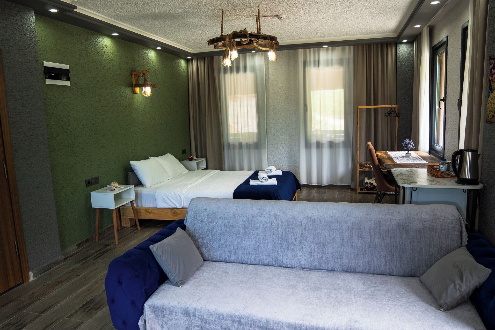 A hotel room with a bed, a sofa, a desk with a chair, a hanging light fixture, and a window with curtains.