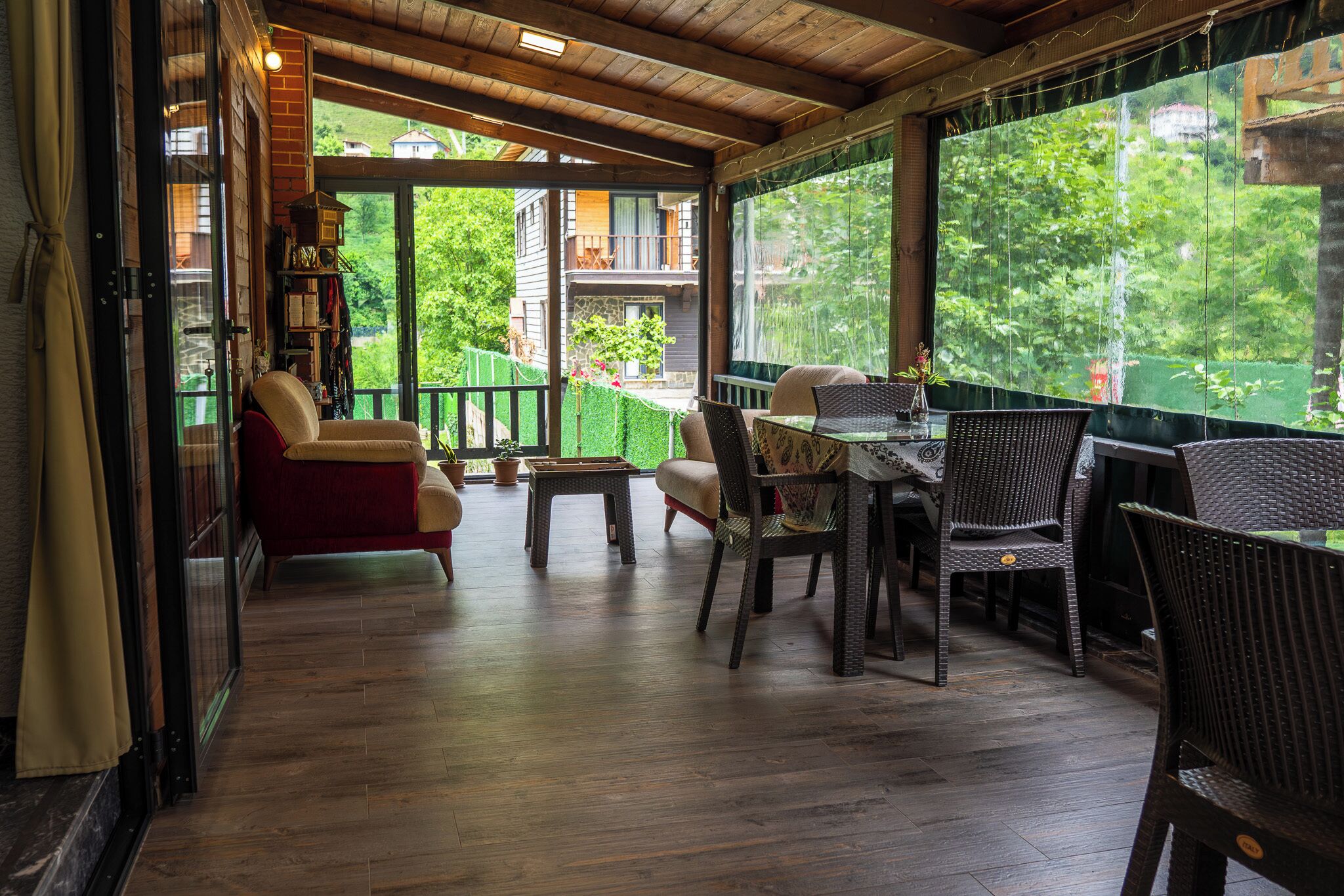 A covered outdoor seating area with wooden flooring, wicker furniture, and a view of a house through large windows.