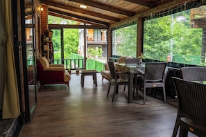 A covered outdoor seating area with wooden flooring, wicker furniture, and a view of a house through large windows. - Pallas Suit  (Ardesen)