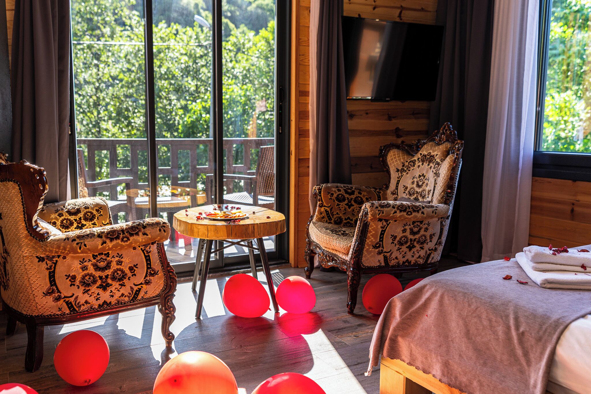 A room with a wooden floor, a round table, two patterned armchairs, a bed with a white cover, and a balcony with a wooden railing.