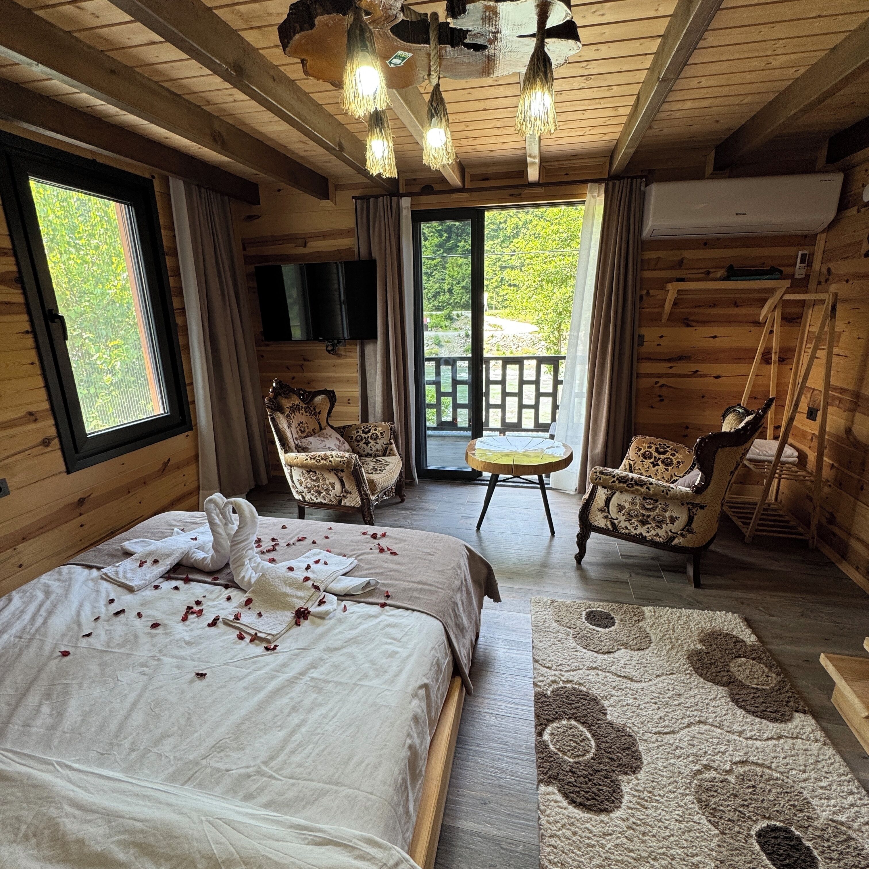 A wooden cabin bedroom with a bed, two chairs, a small table, and a balcony with a view of greenery.