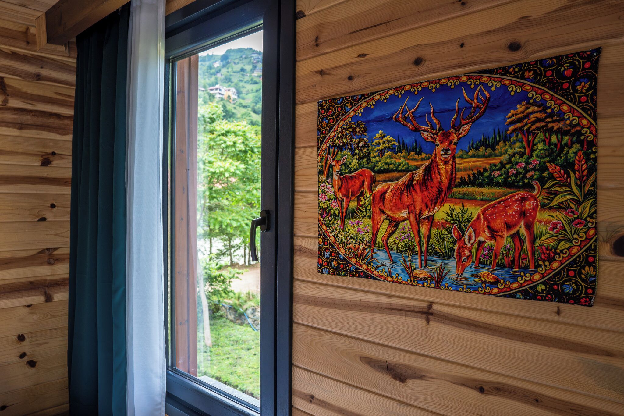 A wooden cabin interior with a window, a tapestry depicting deer, and a view of greenery outside.