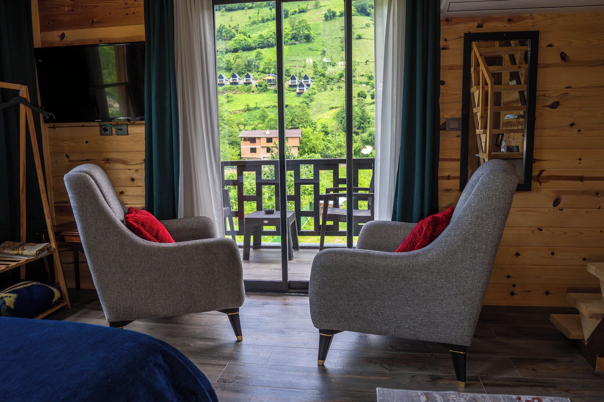A room with two grey armchairs, a wooden TV stand, and a large window with a view of a green landscape.