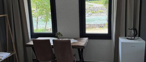 Una habitación con una mesa de madera, dos sillas, un refrigerador pequeño y vistas de un río a través de dos ventanas.