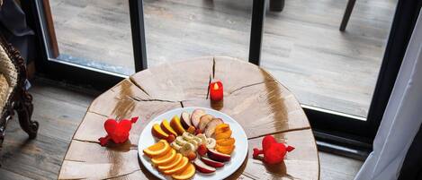 A round wooden table with a plate of fruit and candles on a balcony with a view of a garden and a white vehicle in the background.