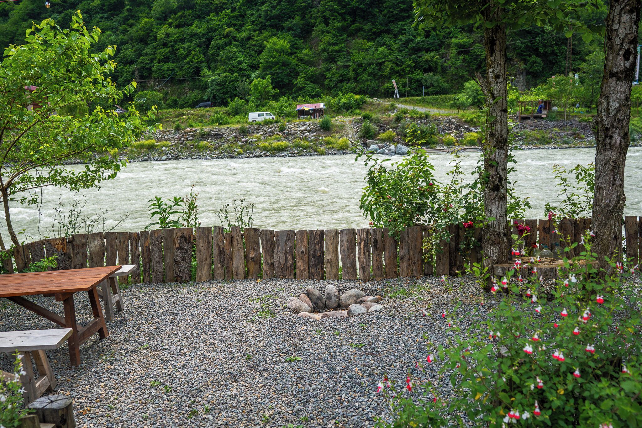 Een camping aan de rivier met een houten omheining, picknicktafels en een vuurkuil.