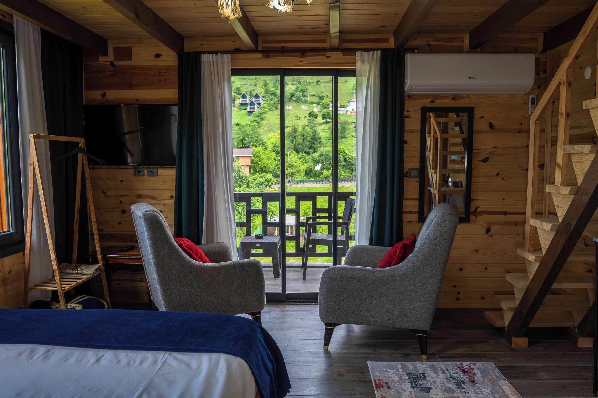 A wooden cabin interior with a bed, two armchairs, a small table, and a staircase.
