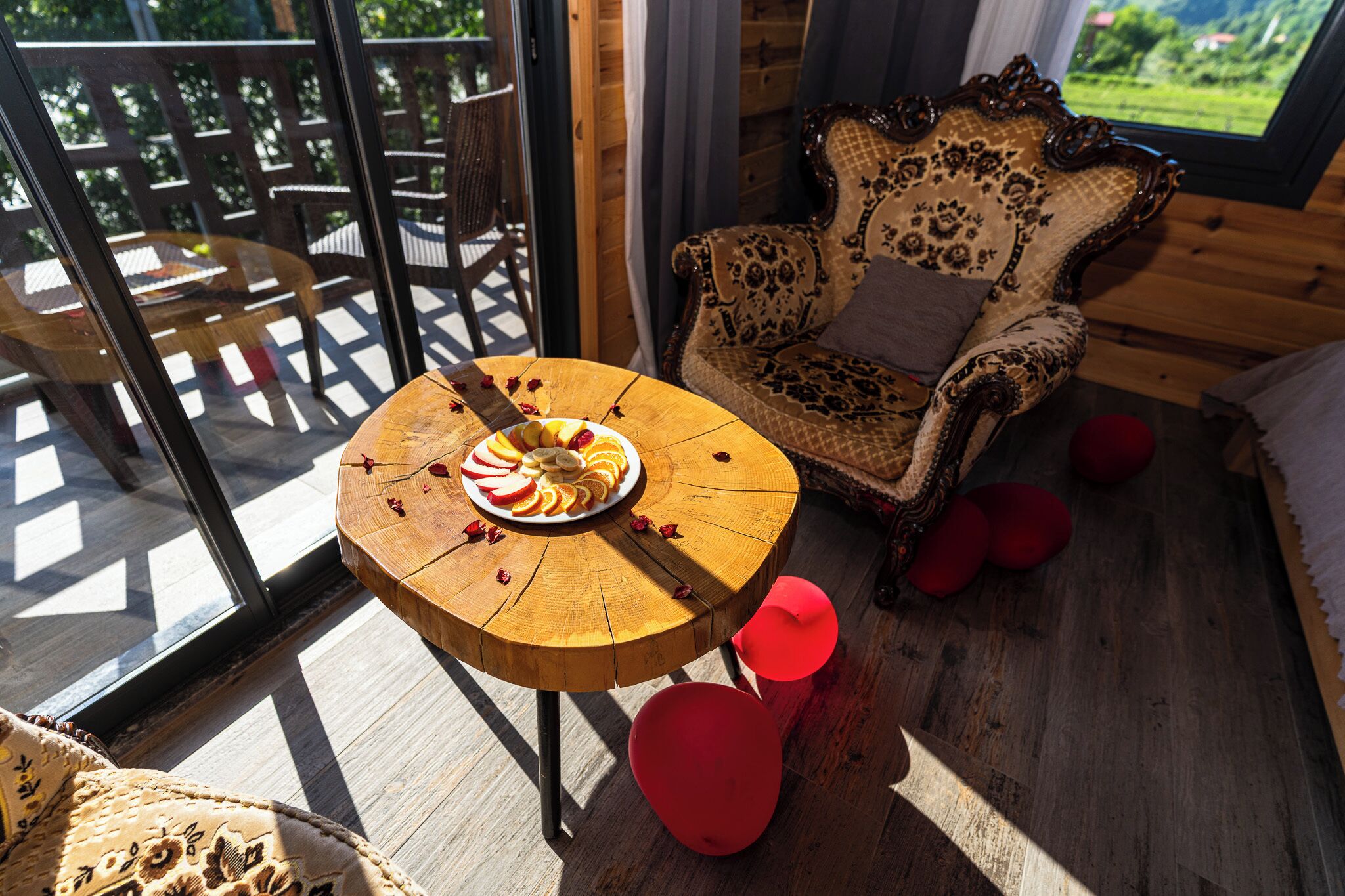 Een houten eettafel met een fruitschaal, een gestreept fauteuil en uitzicht op een houten terras door een glazen deur.