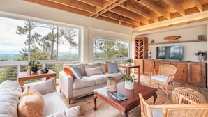 A spacious living room with a high ceiling, large windows, a sofa, a coffee table, wicker chairs, and a television. - Mountain Views 11br 20 People Jarabacoa (Jarabacoa)