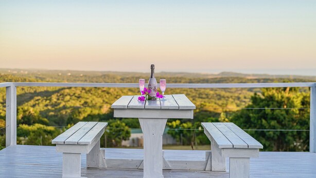 Una mesa de madera blanca con una botella y vasos en un balcón con vistas a un paisaje pintoresco.