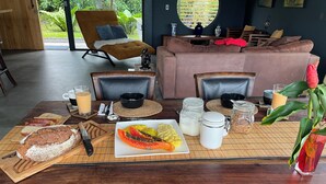 Colazione preparata con pane, frutta e bevande su un tavolo di legno in una stanza con un divano e una poltrona.