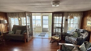 Een ruime woonkamer met houten vloeren, een groene bank, een schommelstoel en uitzicht op zee door grote ramen.