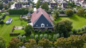 Ett flygbildsbild av ett bostadsområde med ett framträdande hus som har ett brant, spetsigt sadeltak och omges av välskötta gräsmattor, träd och en liten damm.