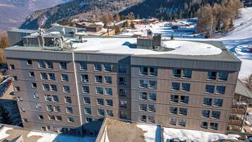 Un hôtel à plusieurs étages, doté d’un toit plat, de grandes fenêtres et offrant une vue sur un paysage enneigé avec des montagnes en arrière-plan.