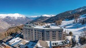 A modern hotel with multiple floors, a rooftop pool, and a ski resort in the background.
