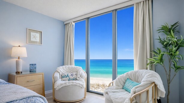 A bedroom with a large window overlooking a beach, two white chairs, a bedside table with a lamp, and a framed picture on the wall.